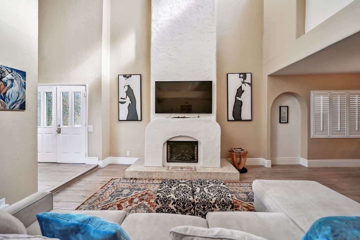 A closer view of the living room's focal point, a tall white stucco fireplace with a mounted television above it. A patterned ottoman and a large sectional sofa rest on a vibrant area rug, complementing the light wood-look flooring.