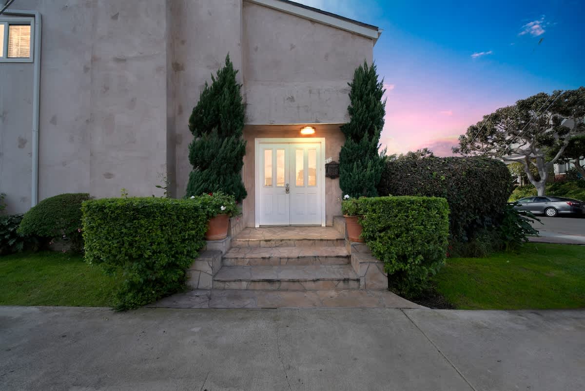 Front exterior of the house featuring a double white entry door flanked by tall evergreen shrubs and manicured hedges, with stone steps leading up to the entrance.