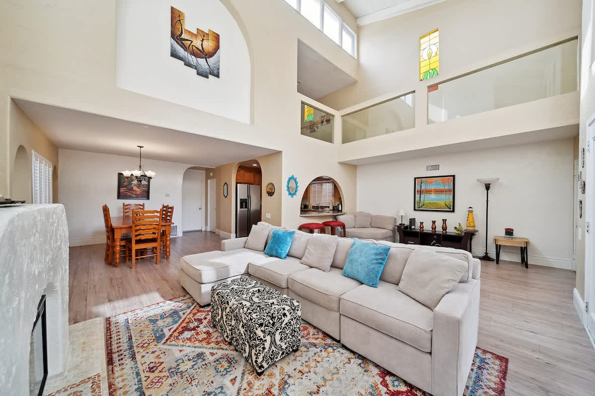 This wide view of the living room showcases a large L-shaped sectional sofa with blue pillows and a patterned ottoman on a colorful area rug. The space features light wood-look flooring, high ceilings, and an open layout towards the dining area and kitchen.