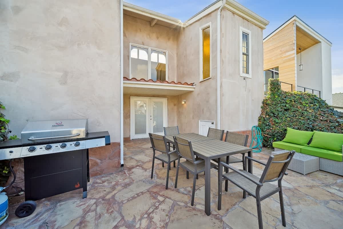 Spacious stone patio featuring an outdoor dining set for six, a BBQ grill, and lush green landscaping, adjacent to the stucco exterior of the house.