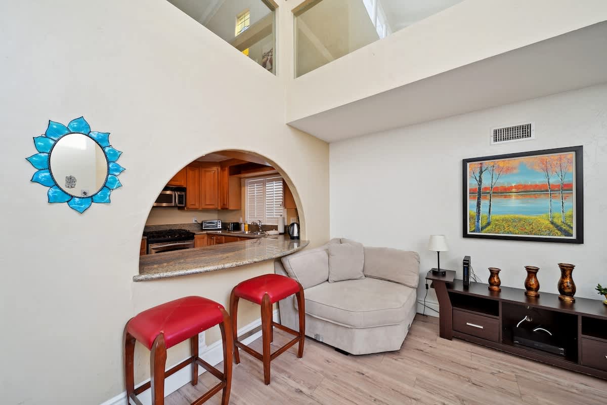 This cozy corner of the living room features a chaise lounge, a dark wood media console, and a large framed painting. An arched pass-through window to the kitchen is visible, complete with two red bar stools.