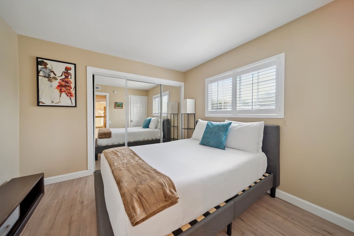 This bedroom features a queen-sized bed with white bedding and a teal accent pillow, light wood flooring, a large mirrored closet, and a window covered by white plantation shutters.