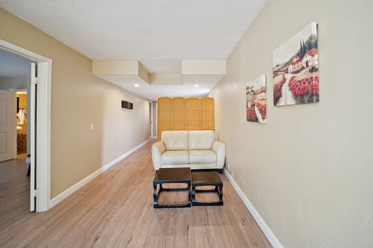 A narrow common area features light wood-look laminate flooring, beige walls, and a cream two-seater sofa paired with two black square coffee tables. A woven room divider stands behind the sofa, while two landscape paintings adorn the right wall, and recessed lighting illuminates the space.