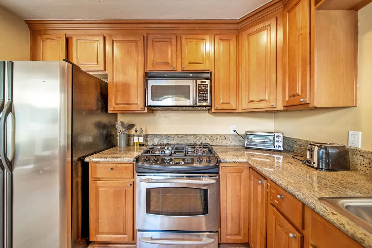 A well-lit kitchen features light wood cabinetry, granite countertops, and stainless steel appliances including a refrigerator, gas range, and microwave, with a toaster and toaster oven on the counter.