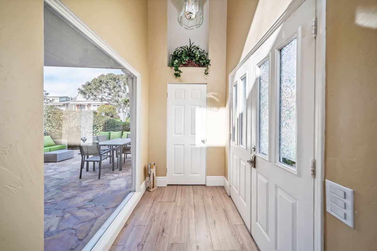 Bright foyer with light wood-look flooring, a white double entry door, and a large glass sliding door offering a view of the outdoor patio and garden.