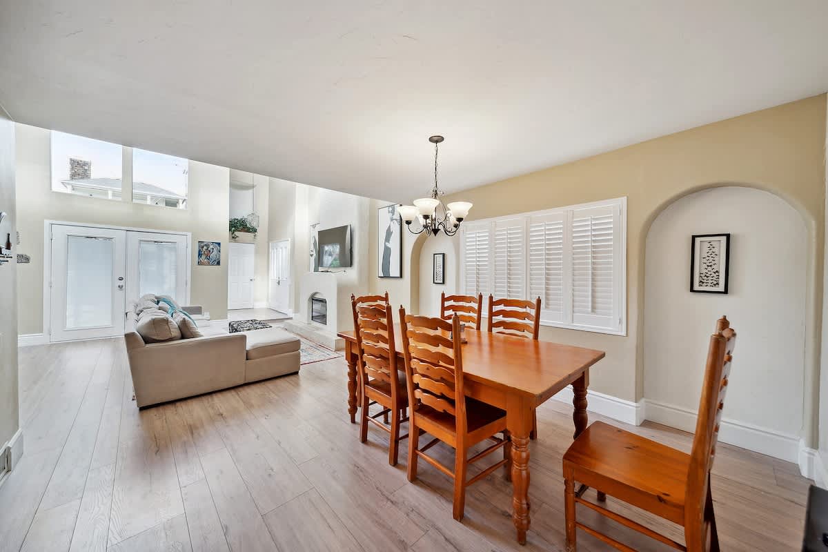An open-concept space features a wooden dining table with six chairs in the foreground, leading to a living room with a light-colored sectional sofa, light wood flooring, and large windows.