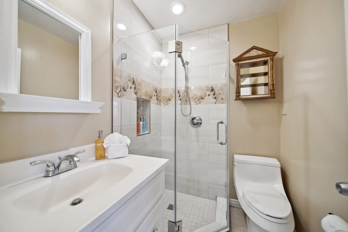 Modern bathroom with a glass-enclosed shower featuring decorative tiled walls, a white vanity with a sink, and a toilet, all set against light beige walls.