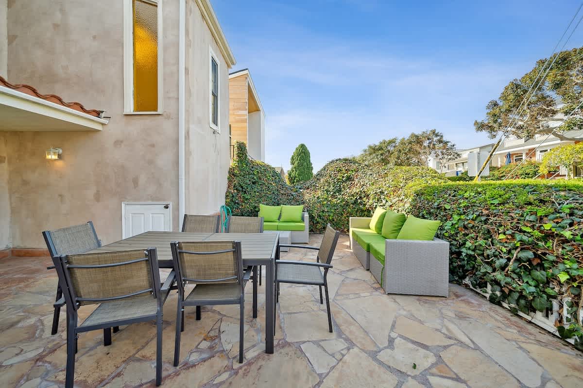 Expansive stone patio with an outdoor dining area and a comfortable lounge seating arrangement with bright green cushions, surrounded by dense green hedges and landscaping.