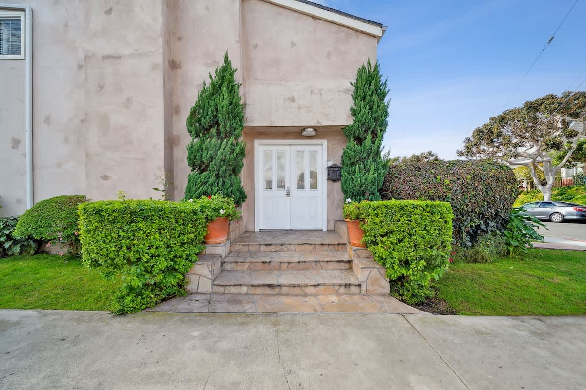 Front exterior of the house showcasing a white double entry door, framed by tall, slender evergreen trees and neatly trimmed hedges, with stone steps leading to the entrance.