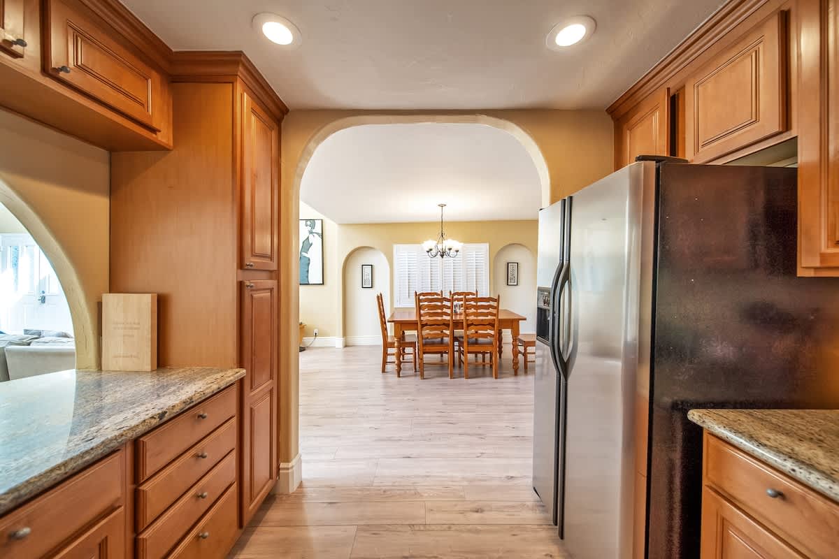 View from the kitchen through an arched doorway into a dining area with a wooden table and chairs, leading to a bright living room with light wood flooring and recessed lighting.