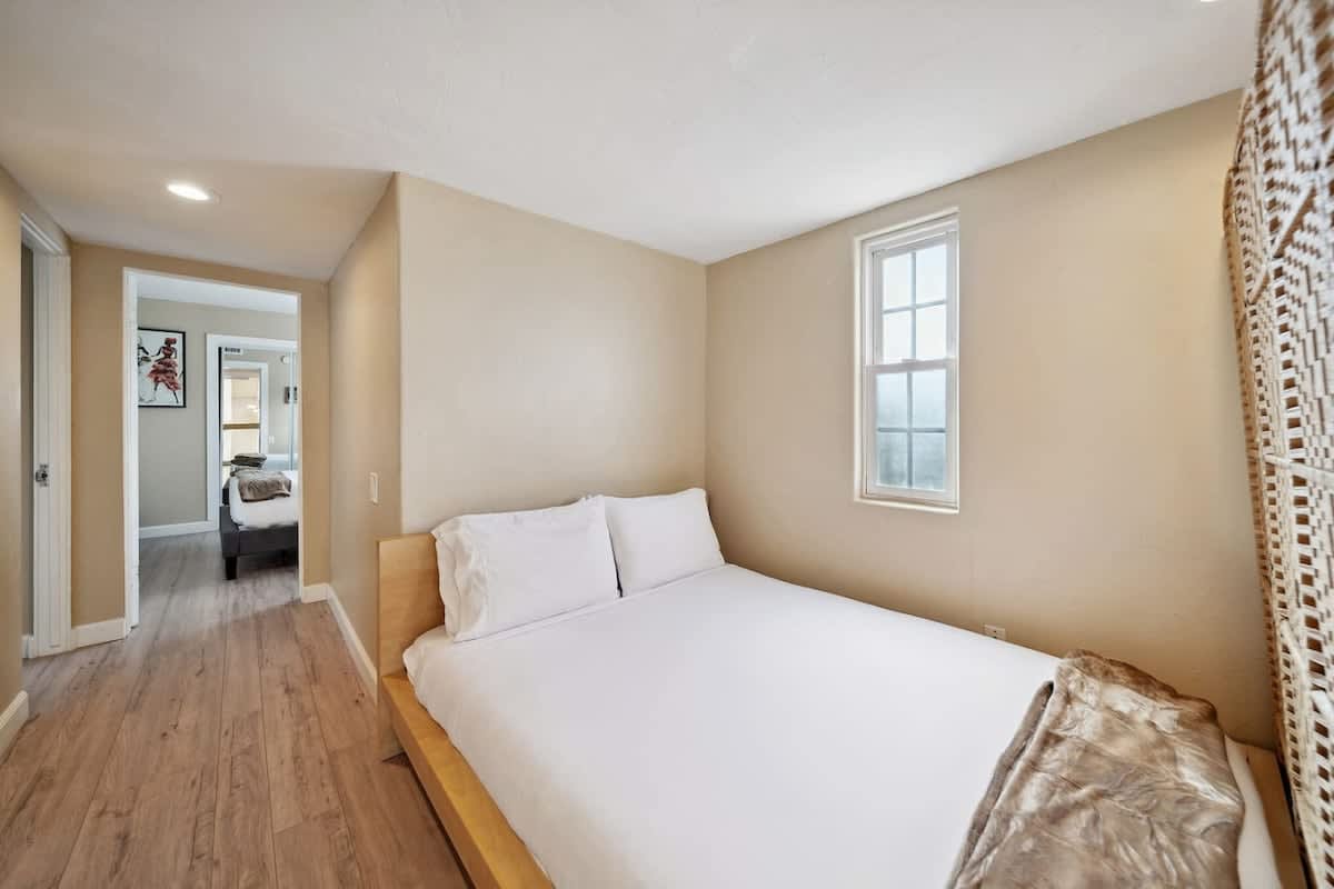 Cozy bedroom featuring a queen-sized bed with crisp white linens, light wood-look flooring, and a small window, with a hallway leading to another room visible in the background.