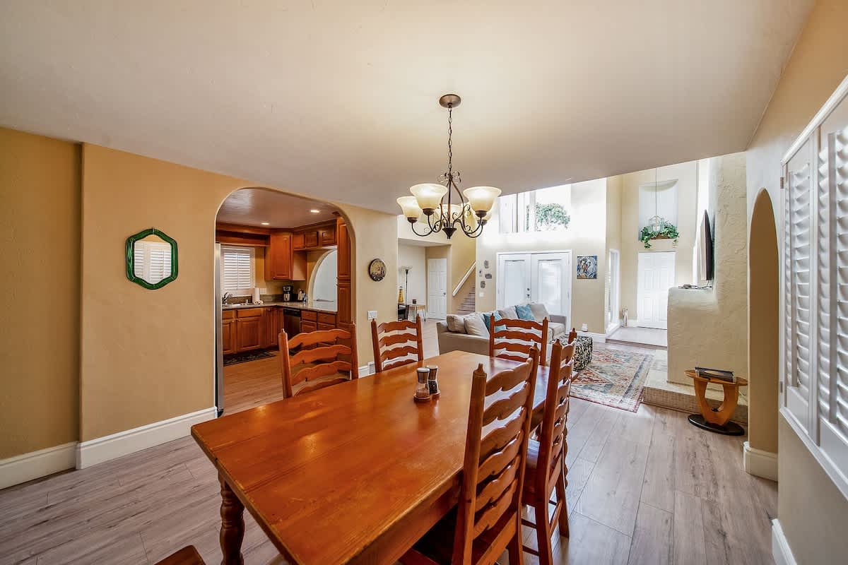 The dining area with a wooden table and chairs is open to a bright living room featuring a fireplace, light wood flooring, and a view towards a multi-level ceiling and large windows.