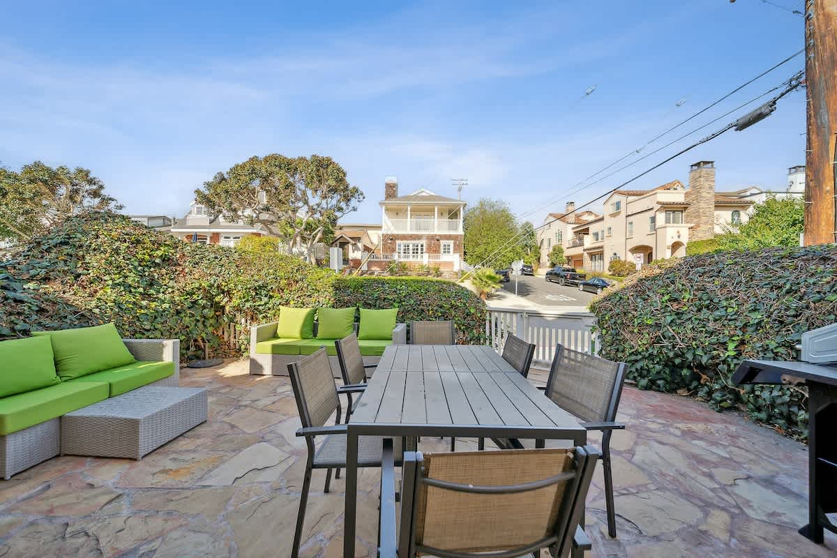 An inviting outdoor patio area with flagstone paving offers a large dining table with chairs and a separate lounge seating area with bright green cushions. Lush green hedges provide privacy, and a BBQ grill is ready for use.
