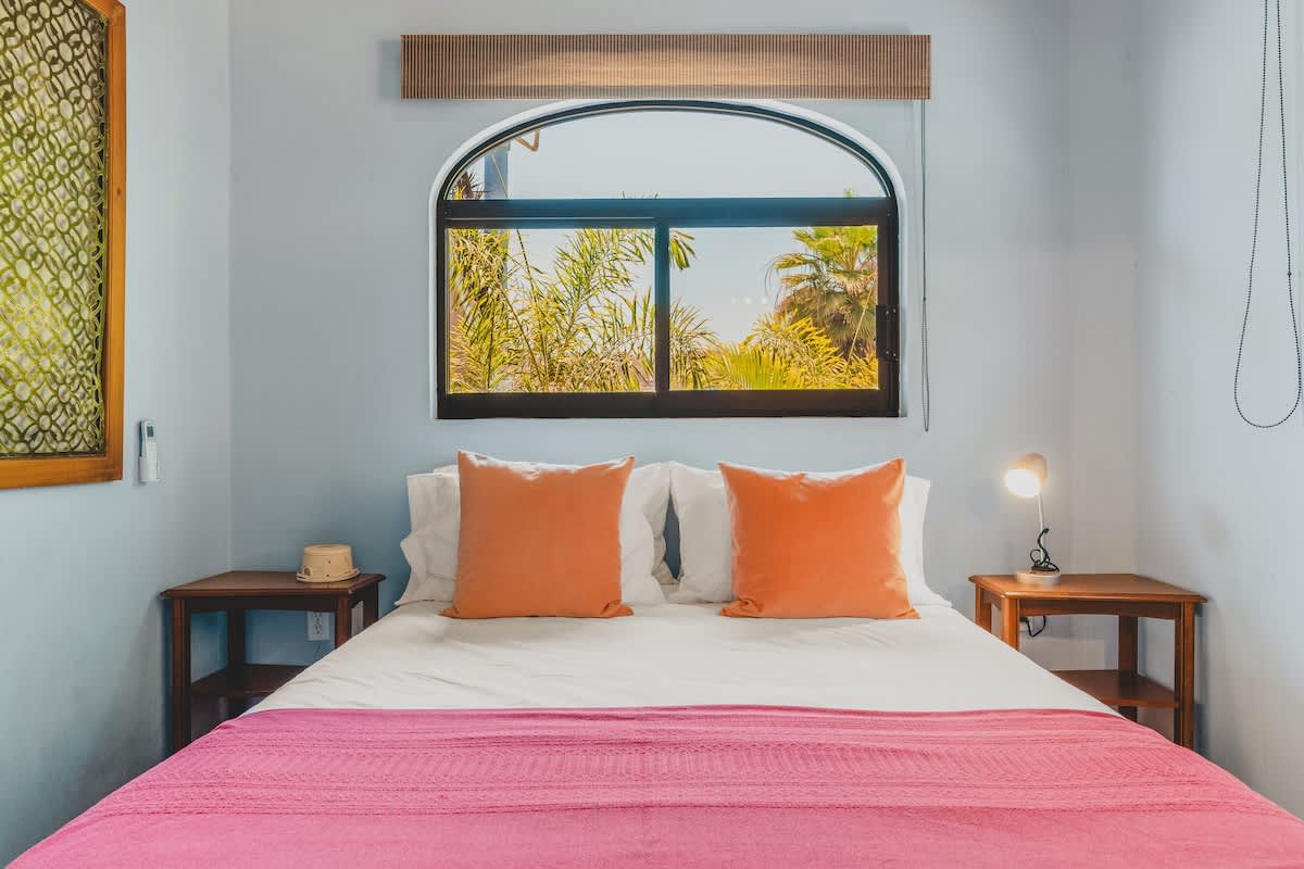 A welcoming bedroom features light blue walls, a queen-sized bed with white linens, orange pillows, and a pink blanket, with wooden nightstands flanking the bed and an arched window providing natural light.