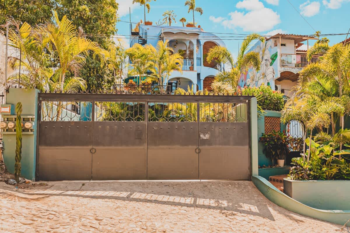 A large, dark metal entrance gate with decorative elements, set into a light blue wall, providing access to a property with lush green foliage and palm trees visible behind it, under a bright sky.