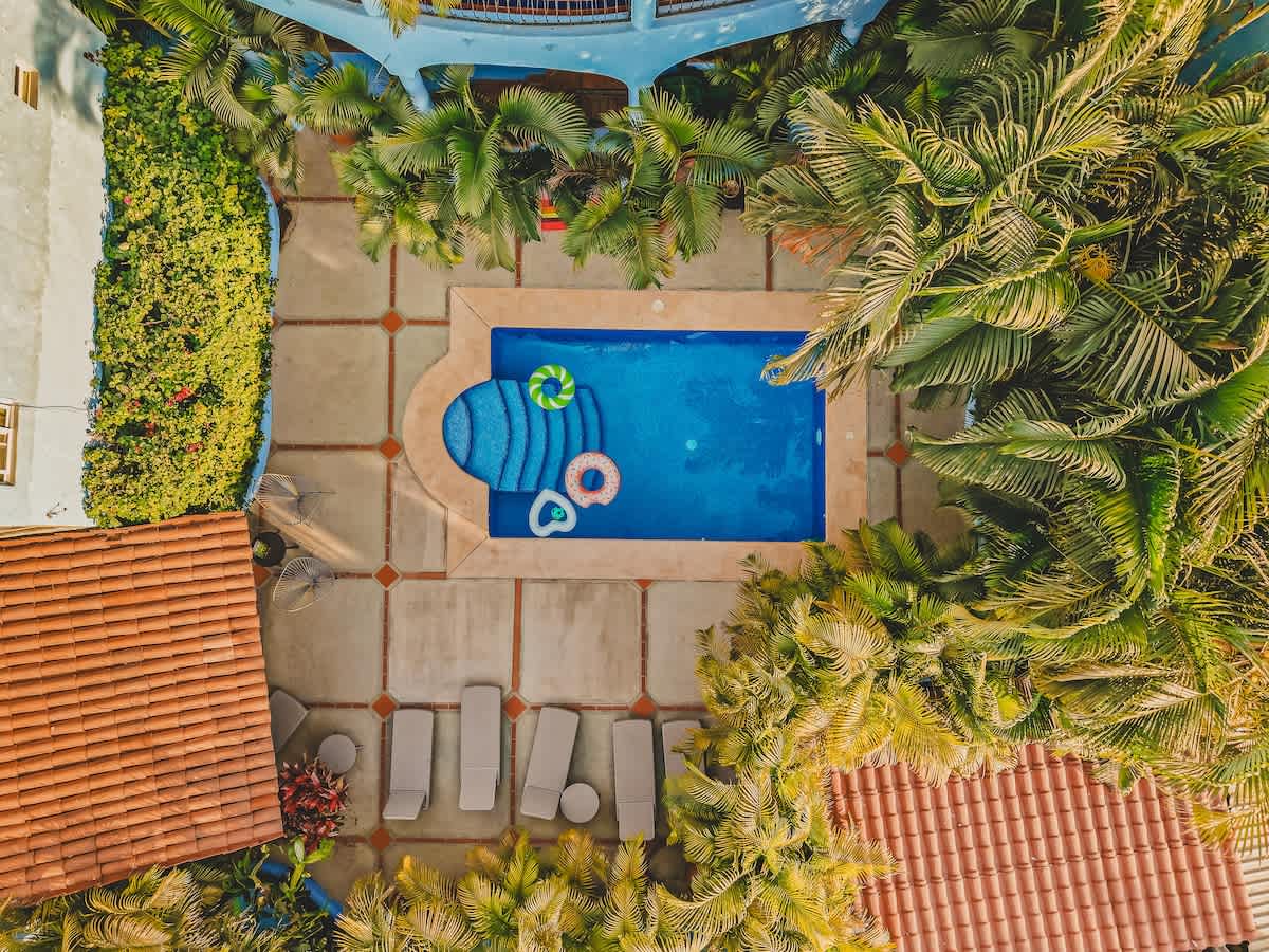 Aerial view of the rectangular swimming pool with blue water and inflatable floats, surrounded by a concrete patio, lounge chairs, and dense tropical foliage.