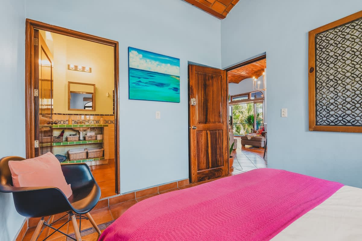A vibrant bedroom features light blue walls, a queen-sized bed with white linens, orange pillows, and a pink blanket, with a terracotta tile floor and an open doorway leading to a bathroom.