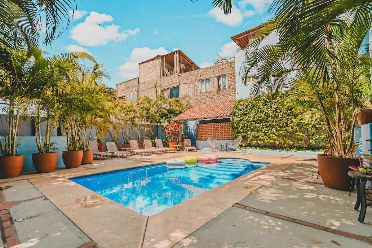 A wide view of a rectangular blue swimming pool with two inflatable rings, surrounded by a spacious concrete patio with multiple lounge chairs, lush palm trees, and a mix of light blue and older buildings under a bright, partly cloudy sky.