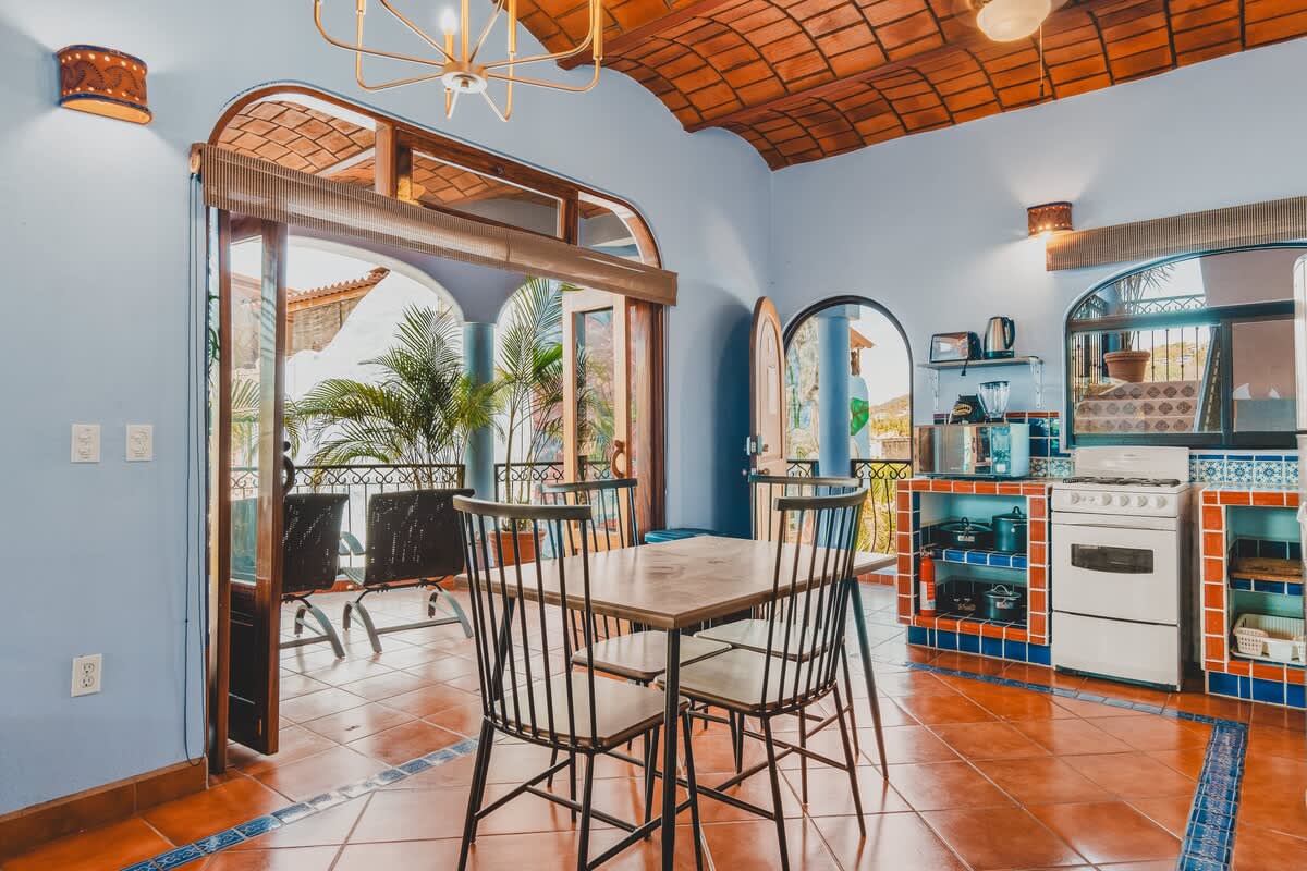 A bright kitchen and dining space with a wooden table and chairs, white stove, and blue-tiled accents, featuring a vaulted terracotta ceiling and large glass doors opening to the balcony.
