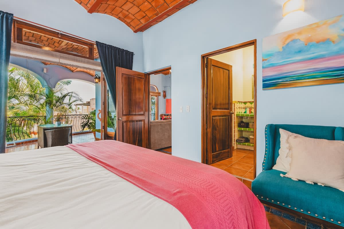 A bedroom with light blue walls and a terracotta vaulted ceiling features a large bed with a red throw blanket. Wooden double doors lead to an adjacent bathroom with green and white tiles, while another set of doors opens to a living area.