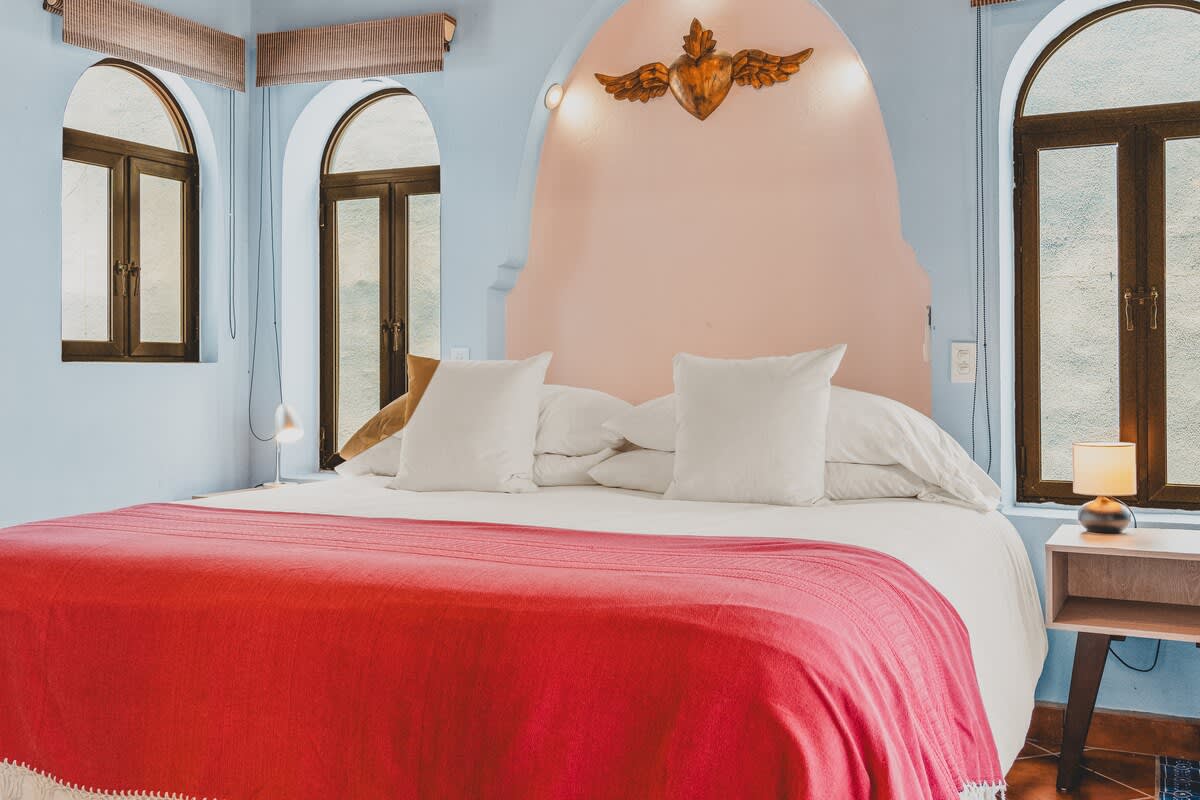 This vibrant bedroom features light blue walls, a large bed with a bright red blanket and white pillows, and a unique pink arched headboard adorned with a golden winged ornament. Two tall arched windows with dark frames are visible on the left, complementing the eclectic style.