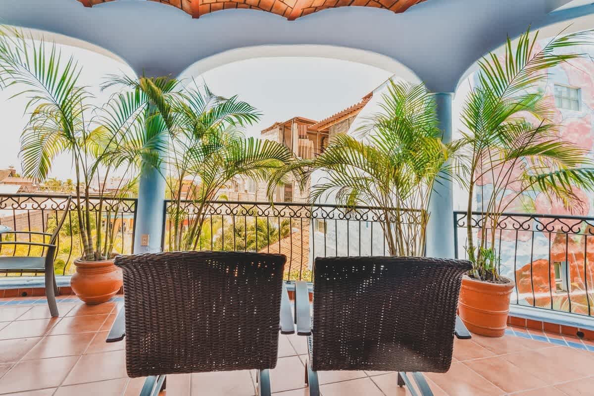 A scenic balcony with two dark wicker chairs, offering views of palm trees and the surrounding neighborhood buildings under a bright sky.