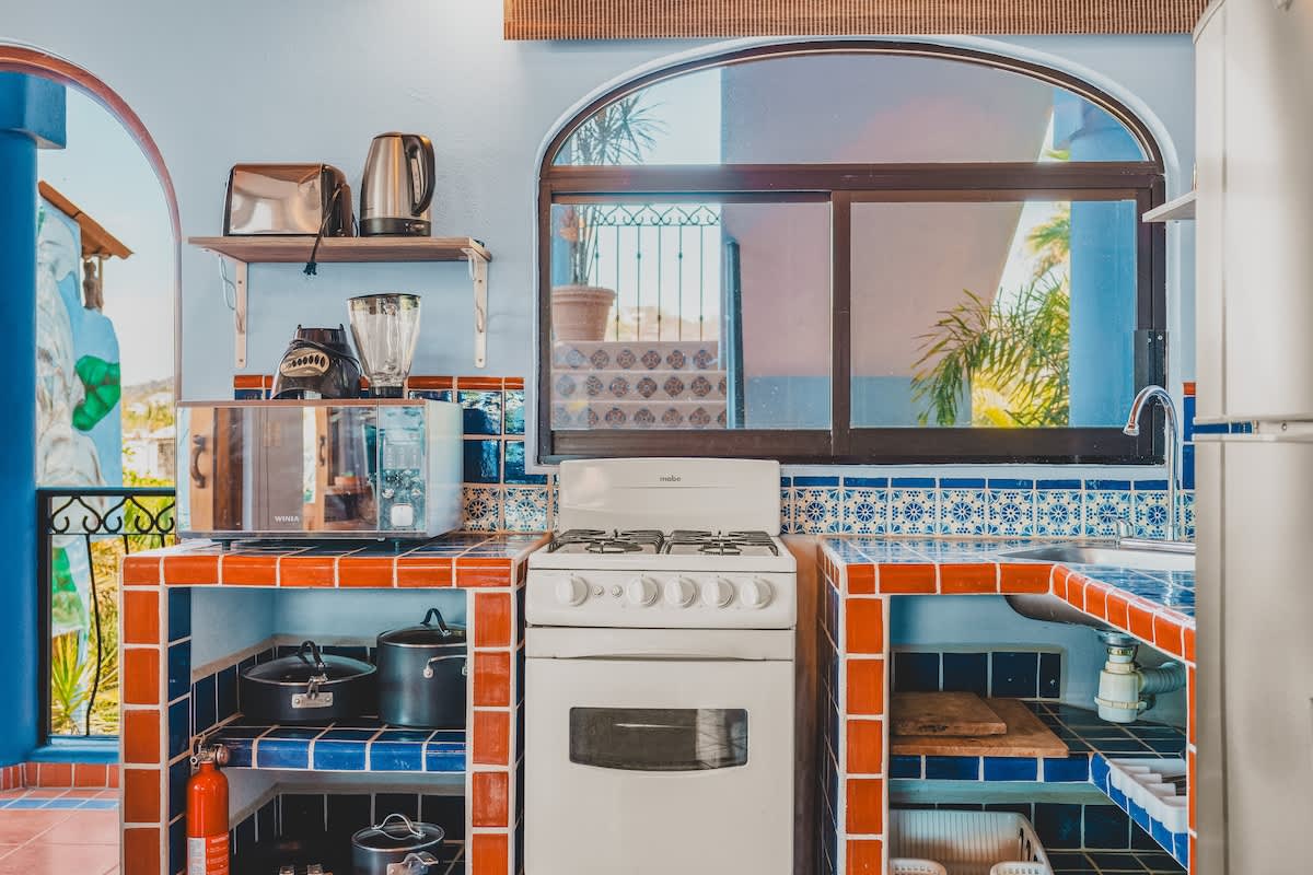A vibrant kitchen with a white gas stove, microwave, blender, and tiled countertops in blue and orange, featuring an arched window and open shelving with pots and pans.