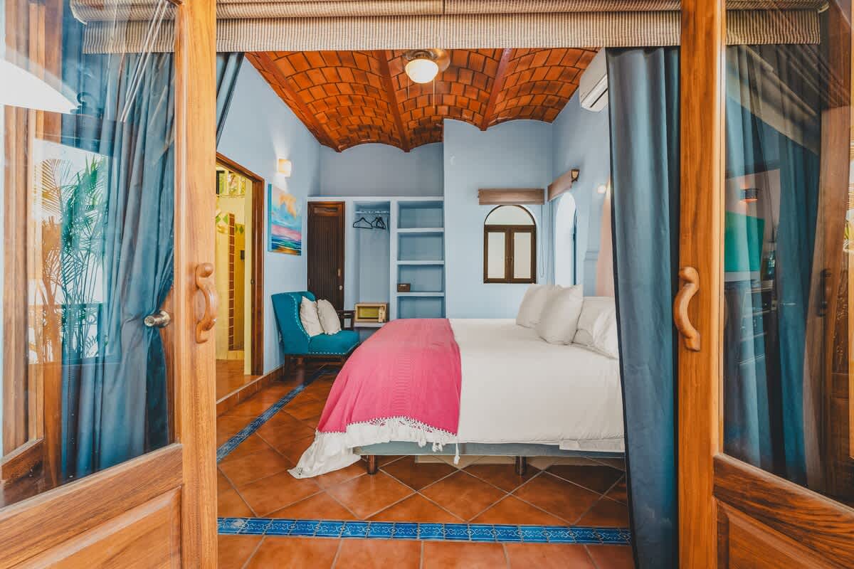 A cozy bedroom with a distinctive terracotta tile ceiling and blue tiled floors, featuring a large bed with a pink blanket and white pillows. The room includes an open closet area, a blue armchair, and a small window, all viewed through open wooden doors framed by blue curtains.