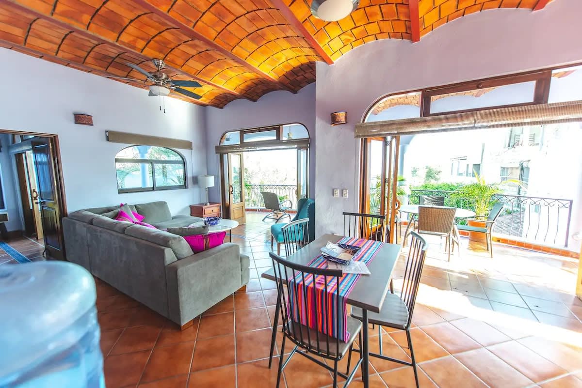 A spacious open-concept living and dining area features terracotta tile flooring and a vaulted terracotta tile ceiling with a ceiling fan. A long grey sofa with colorful pillows faces a dining table with striped placemats, with access to a large balcony.