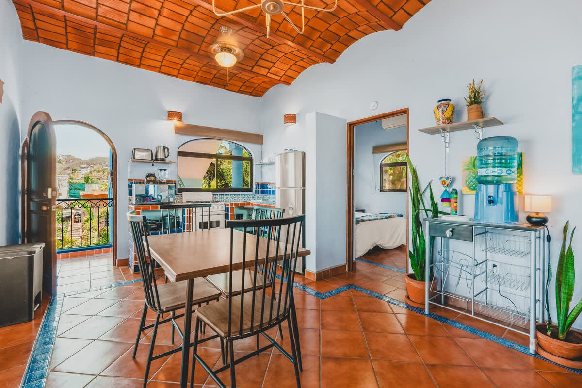 A functional kitchen and dining area with terracotta tile floors, a wooden dining table for four, white appliances, and blue and white tiled countertops, with a bedroom visible through an open doorway.