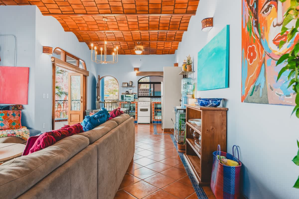 Open-concept living area with a long beige sofa, light blue walls, and a vaulted terracotta tile ceiling, leading to a kitchen area and featuring colorful artwork and patterned tile flooring.
