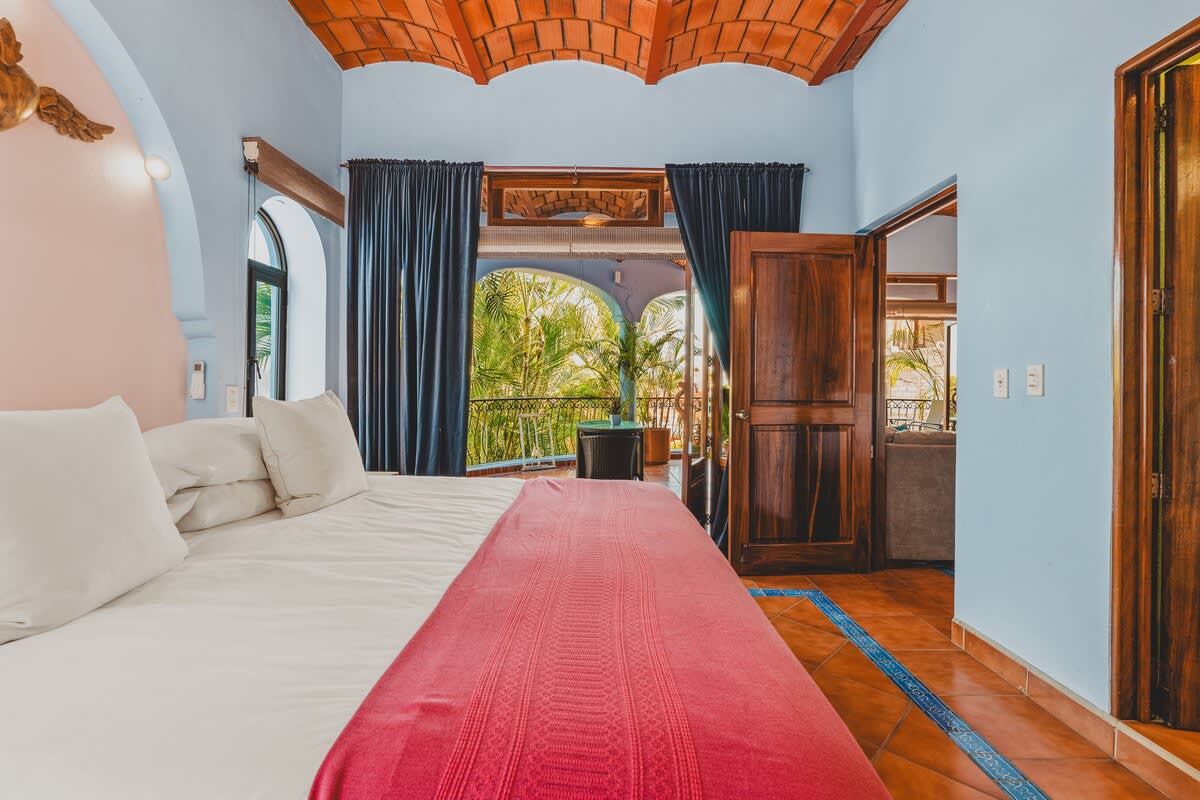 Bright bedroom with a large bed featuring white linens and a pink throw, light blue walls, a vaulted terracotta tile ceiling, and a balcony with lush green views through open doors and dark curtains.
