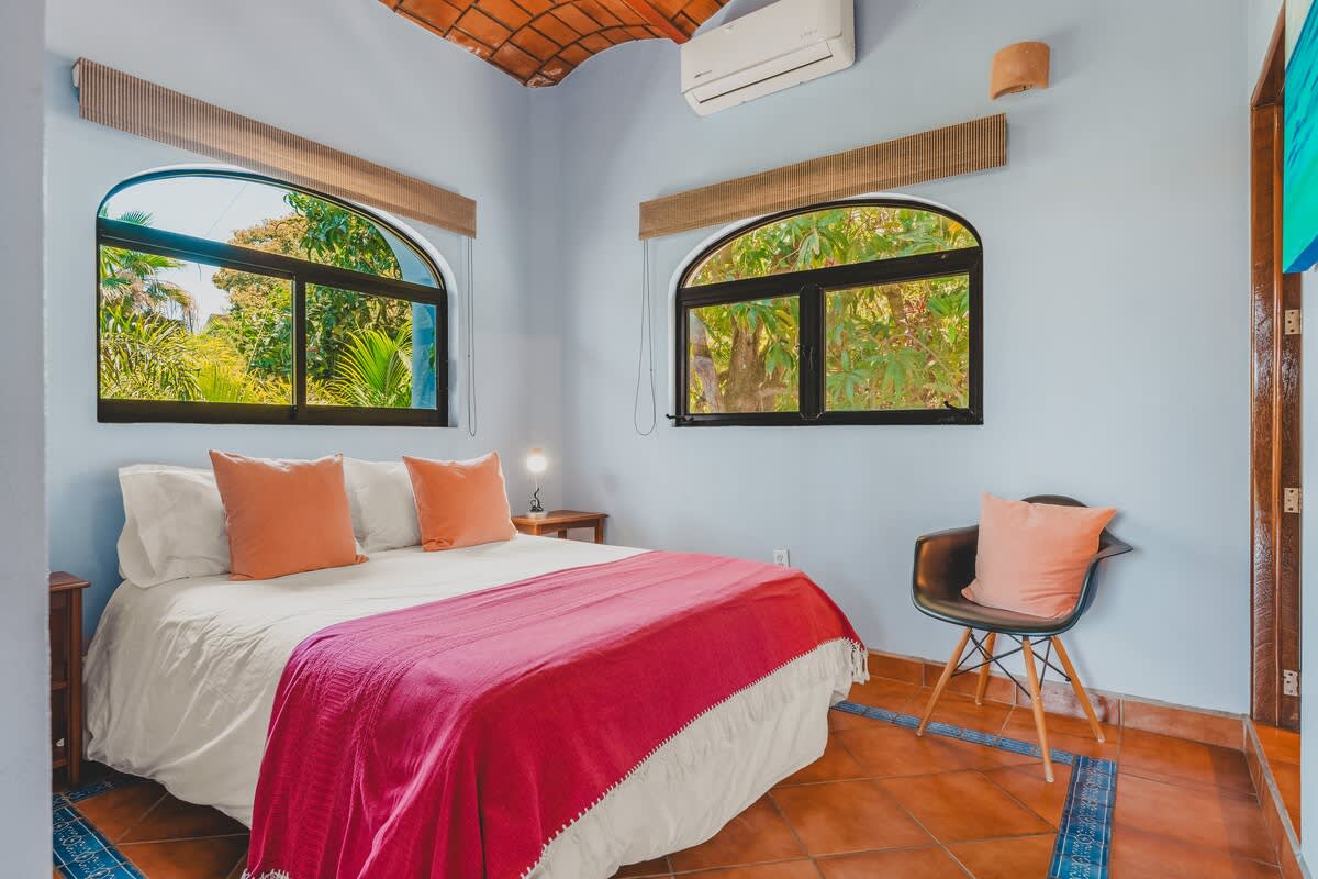 This bright bedroom features light blue walls, a queen-sized bed with white linens, orange pillows, and a pink blanket, complemented by terracotta tile flooring and two arched windows offering views of tropical trees.