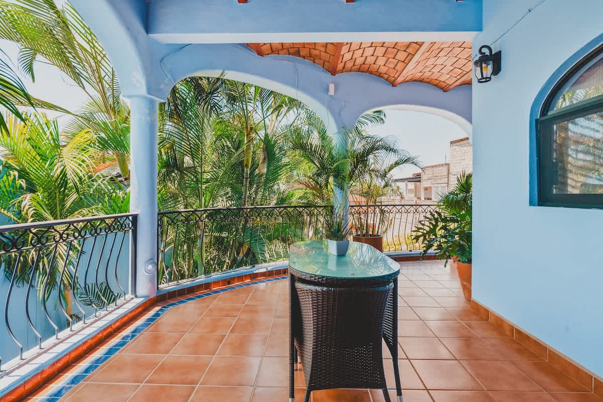A spacious outdoor balcony features terracotta floor tiles and light blue walls, with a terracotta vaulted ceiling. A small round glass-top table with a wicker base and chair is positioned near the wrought iron railing, offering views of lush greenery and distant buildings.
