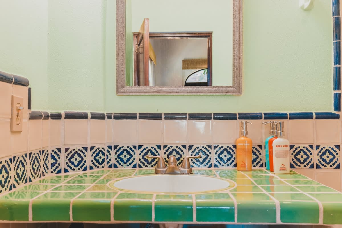 A detailed view of the bathroom sink area highlights a green tiled countertop with a white basin, surrounded by light peach tiles with a blue and white patterned border, and a light green wall with a mirror.