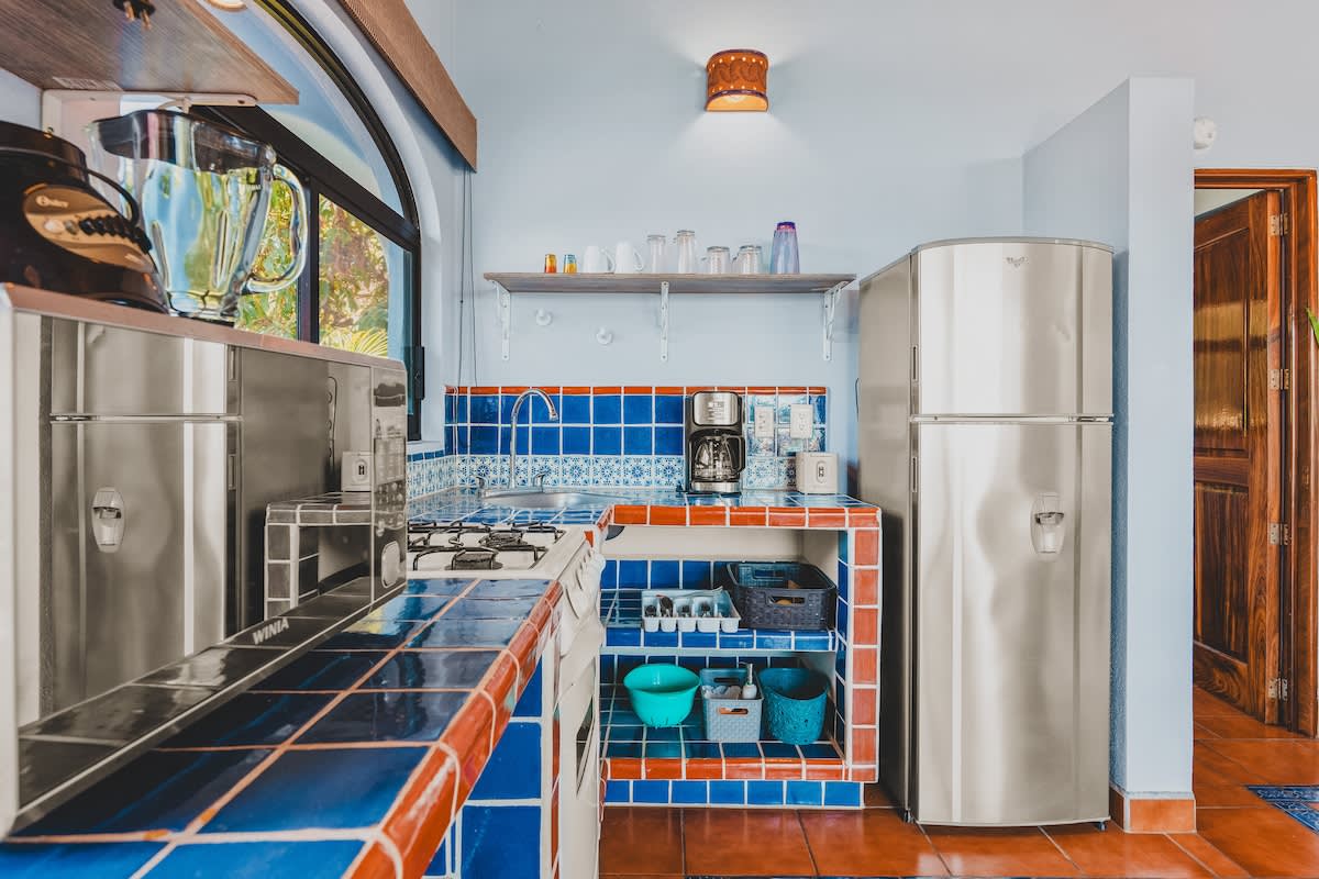 A bright kitchen featuring blue and orange tiled countertops, a stainless steel refrigerator, a sink, and an arched window, with various kitchen appliances and utensils visible.