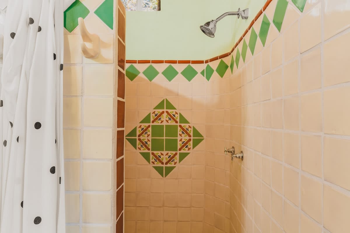 A vibrant shower stall features light beige square tiles with a decorative green and brown diamond pattern border and a central large decorative tile. The walls above the tile are light green, and a white polka-dotted shower curtain is visible on the left.
