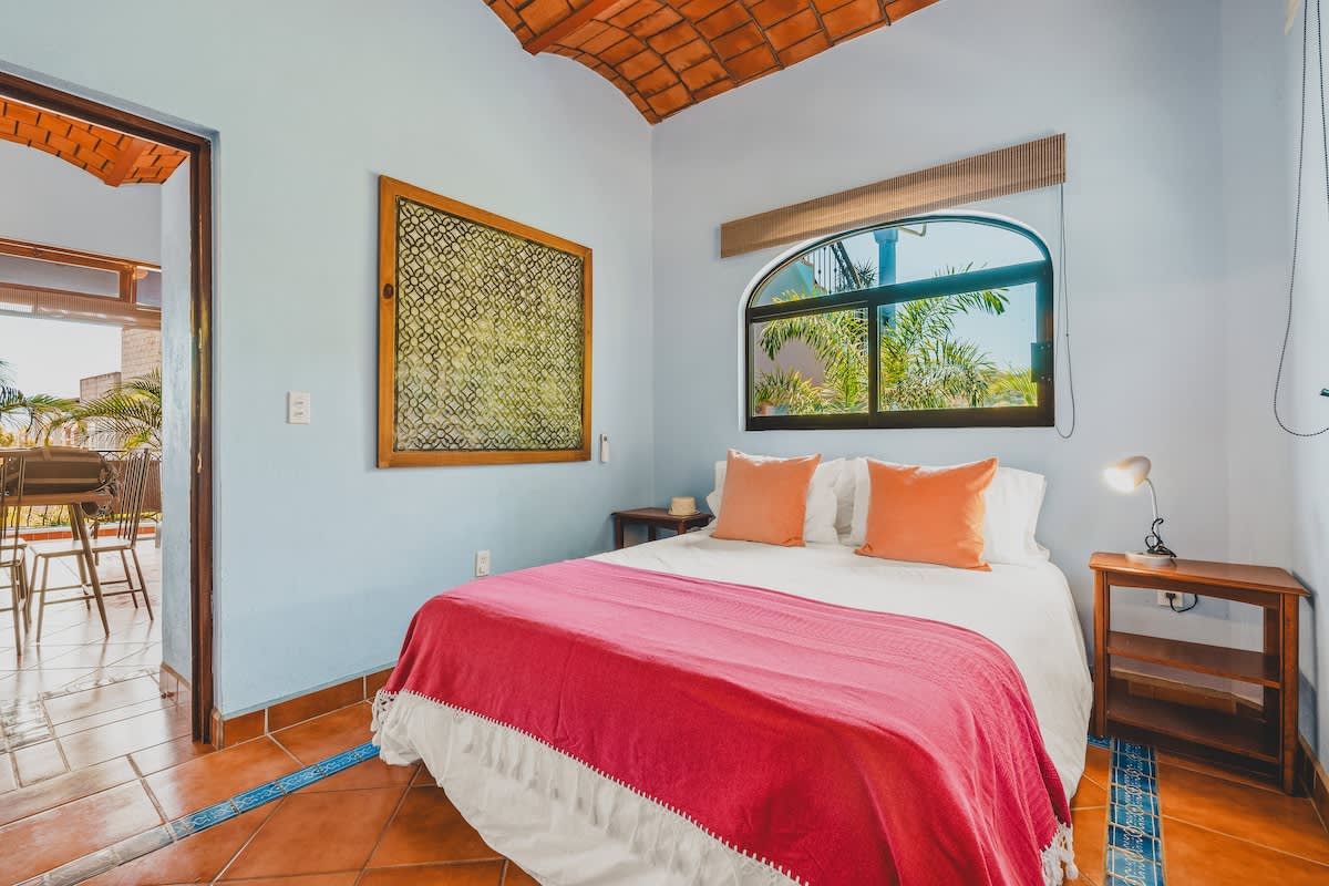 This bright bedroom features light blue walls, a queen-sized bed with white linens, orange pillows, and a pink blanket, with a terracotta tile floor and an arched window offering views of lush trees.