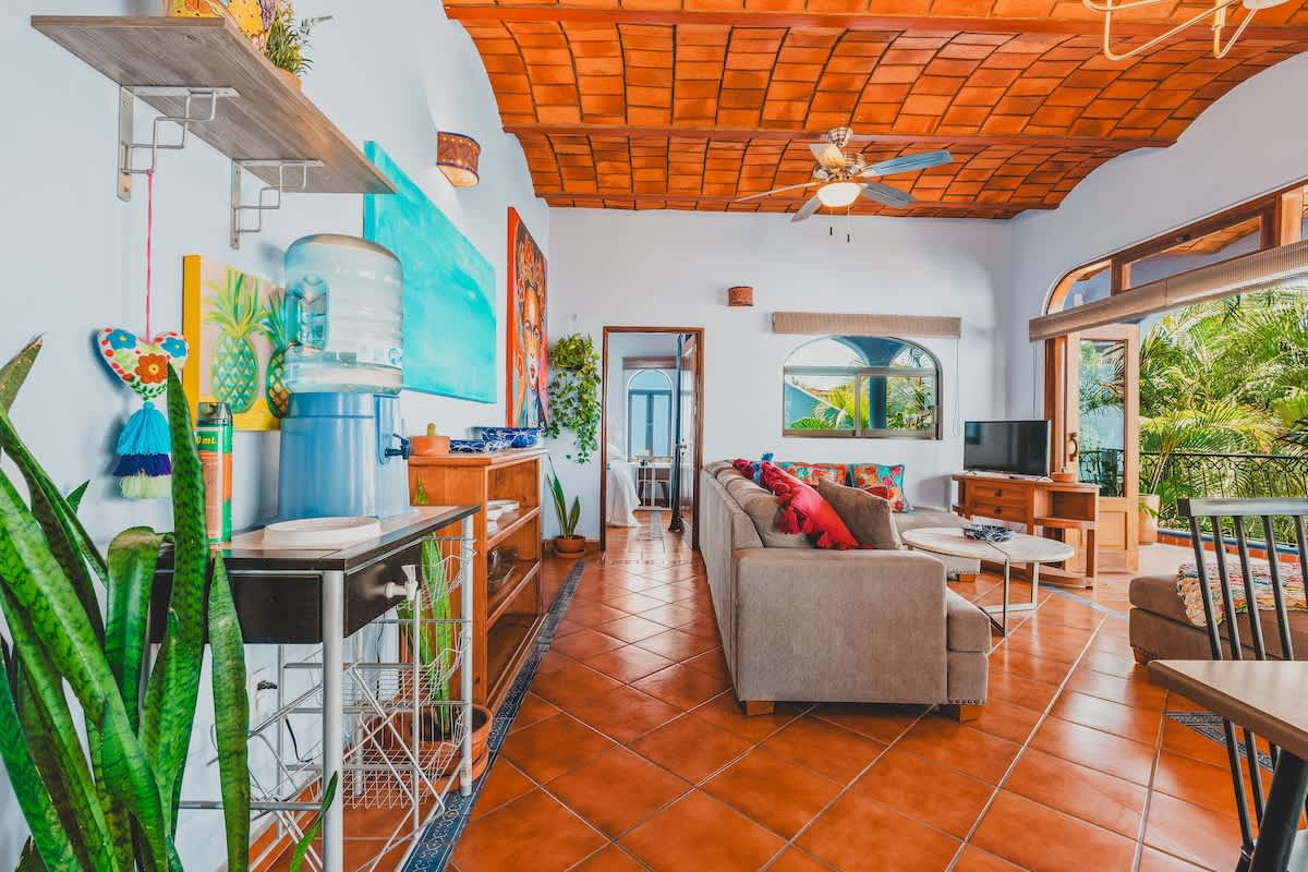 A bright and airy living room with a large L-shaped sofa, a coffee table, a TV, and a water dispenser, featuring a terracotta tiled ceiling and light blue walls.