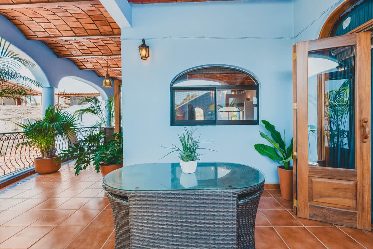 A spacious outdoor balcony features light blue walls, terracotta floor tiles, and a terracotta vaulted ceiling. A glass-top table with a wicker base is positioned on the left, with lush green plants in terracotta pots and a wrought iron railing providing a tropical ambiance.