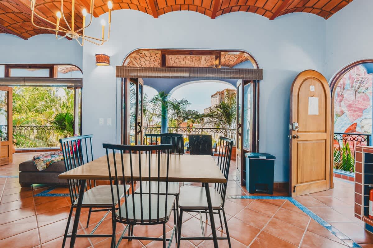 A spacious dining area with a wooden table and black chairs, terracotta tile flooring with blue accents, and a vaulted ceiling, offering views of the balcony through open doors and showing a wooden arched door.