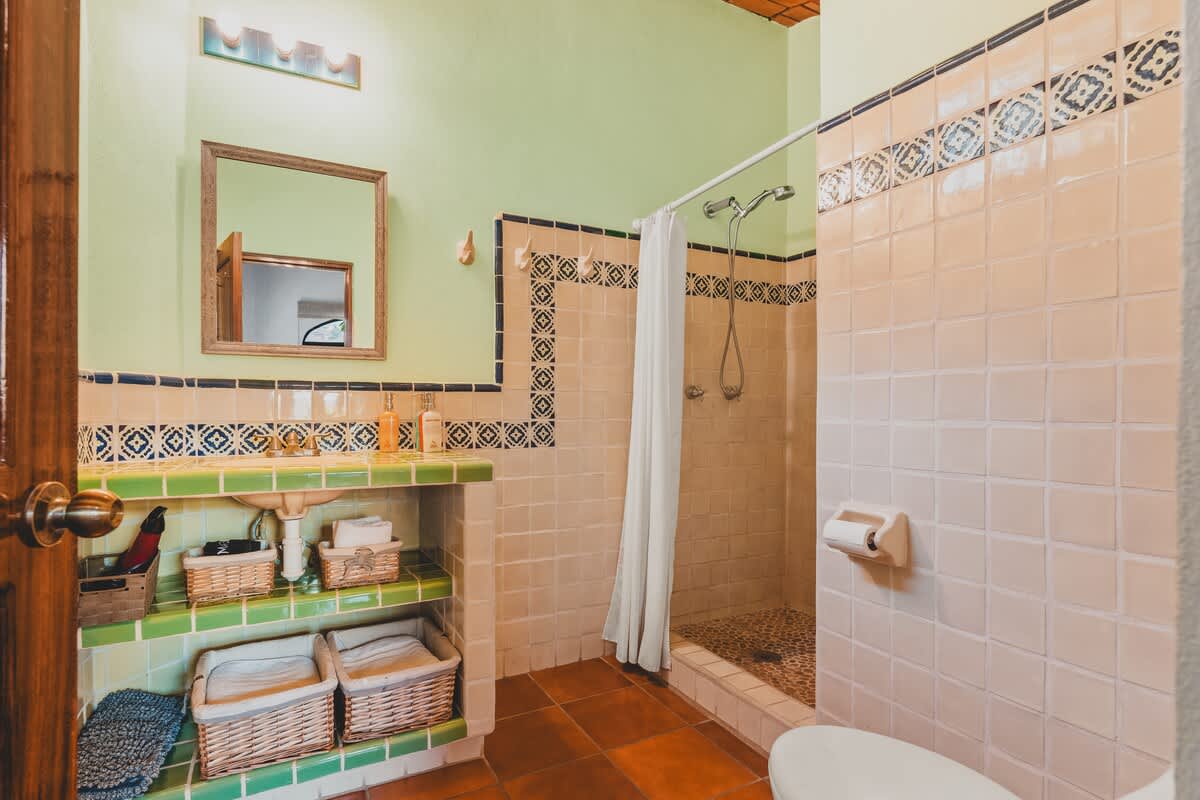 A colorful bathroom features light peach square tiles on the walls with a blue and white patterned border, a green tiled vanity with open shelving, a walk-in shower, and a toilet, all under light green walls.