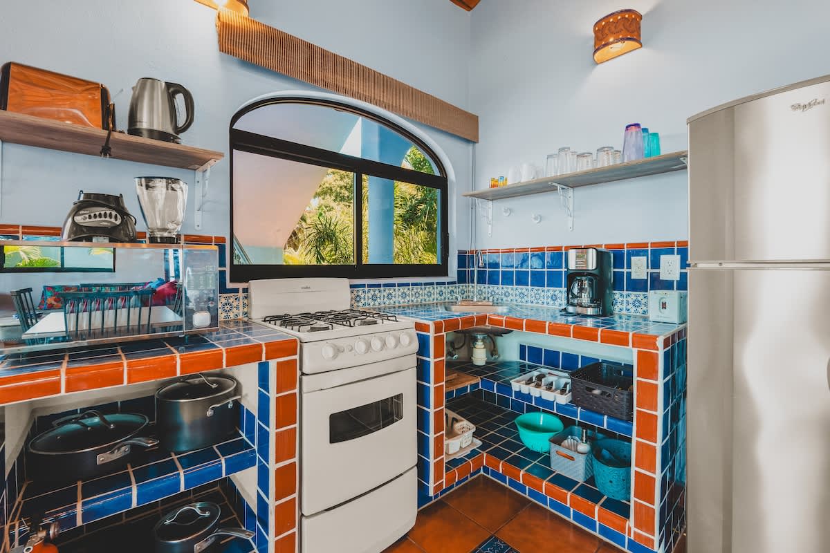 A colorful kitchen with blue and orange tiled countertops and backsplash, a white gas stove, a stainless steel refrigerator, and an arched window overlooking greenery.