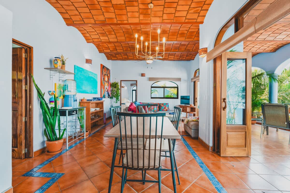 A vibrant dining area with a long wooden table and black chairs, featuring a terracotta tiled ceiling, light blue walls, and a view into a living space and outdoor patio.