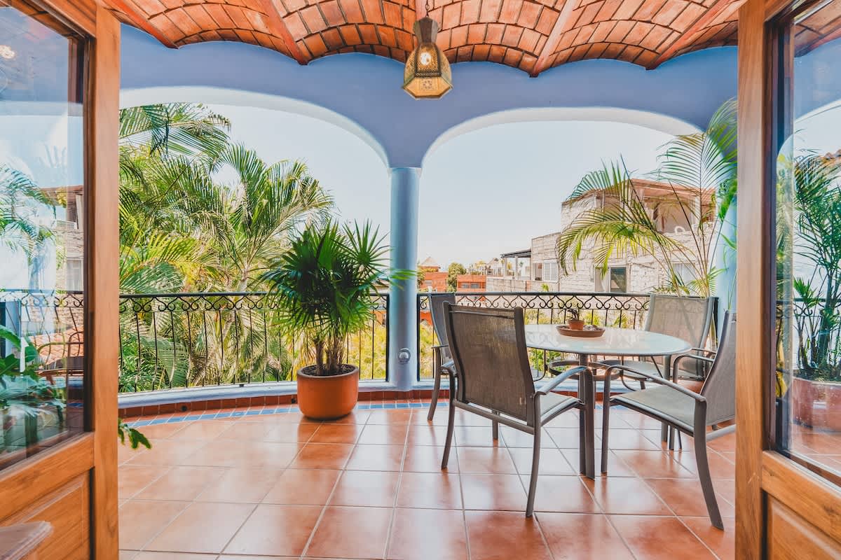 A spacious outdoor balcony with terracotta tile flooring, a glass-top table, and four chairs, surrounded by lush palm trees and offering views of distant buildings.