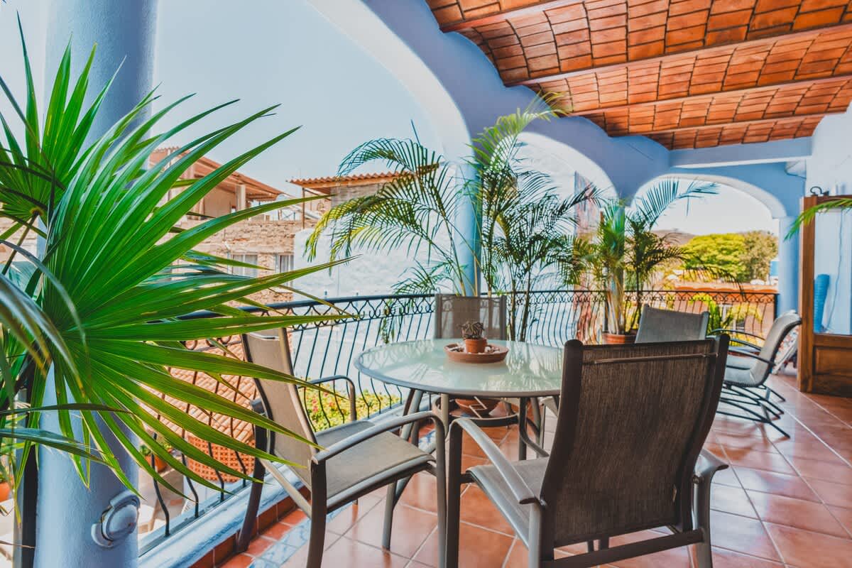 A close-up of the balcony's outdoor dining area, featuring a glass-top table and chairs amidst lush palm foliage, with a glimpse of distant buildings under a clear sky.