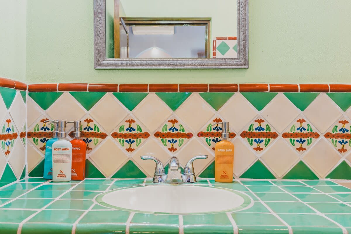 A bathroom vanity features a white sink set into a green tiled countertop, with a decorative border of green, white, and floral patterned tiles above. Various body care products are neatly arranged on the counter, and a simple mirror hangs above.