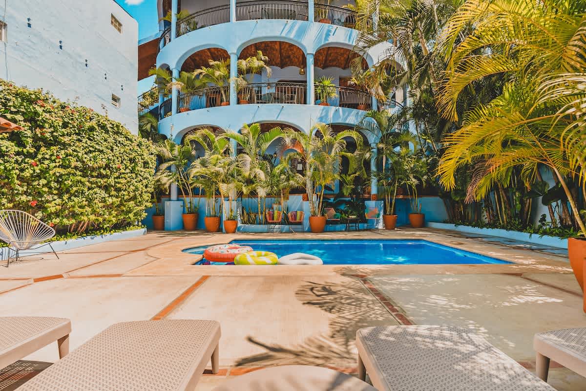 A low-angle view of a rectangular blue swimming pool with two inflatable rings, surrounded by a light-colored concrete patio with lounge chairs, lush palm trees, and a distinctive light blue multi-story building with arched balconies.