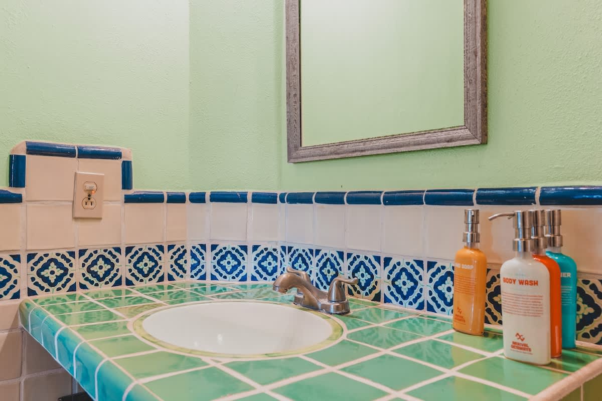 A close-up of the bathroom vanity shows a green tiled countertop with a white sink, surrounded by light peach tiles with a blue and white patterned border, and a light green wall above.