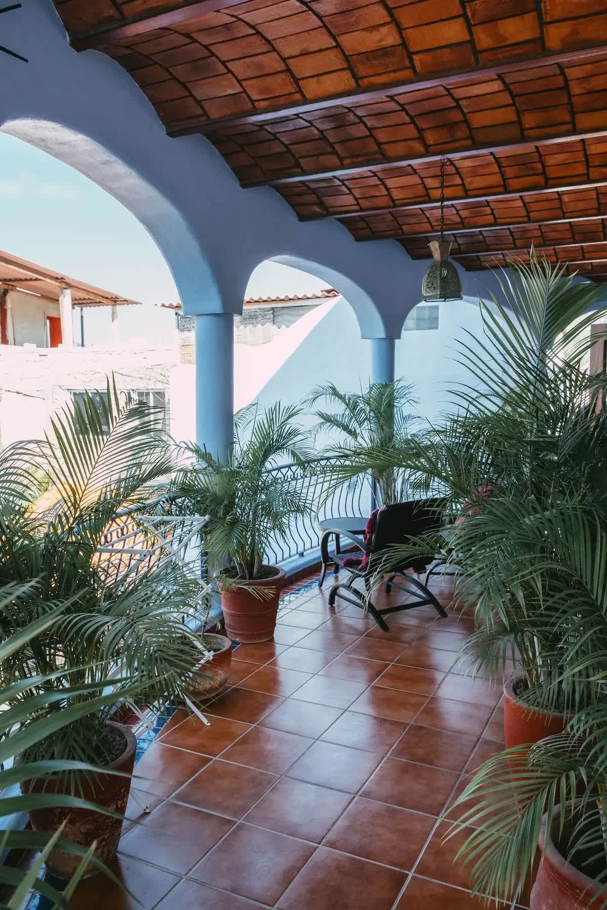 A covered balcony with terracotta tile flooring and light blue arched columns offers outdoor seating with two chairs and a small table. Potted palm trees line the wrought iron railing, providing a tropical ambiance.